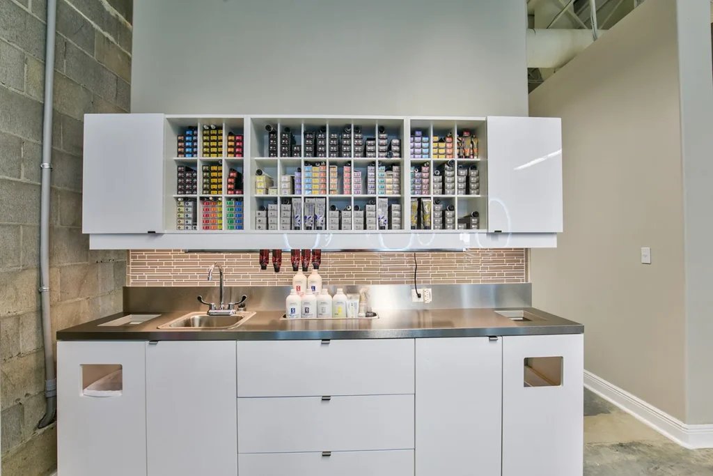 A modern workspace with white cabinets, a stainless steel countertop, a small sink, and shelves neatly stocked with colorful supplies and bottles. - Soleil 7 Hair Salon in Tallahassee, FL