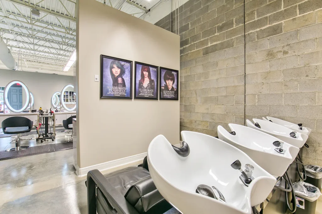 Three salon shampoo stations beside a concrete wall with three framed hairstyle posters; styling chairs and mirrors are visible in the adjoining room. - Soleil 7 Hair Salon in Tallahassee, FL