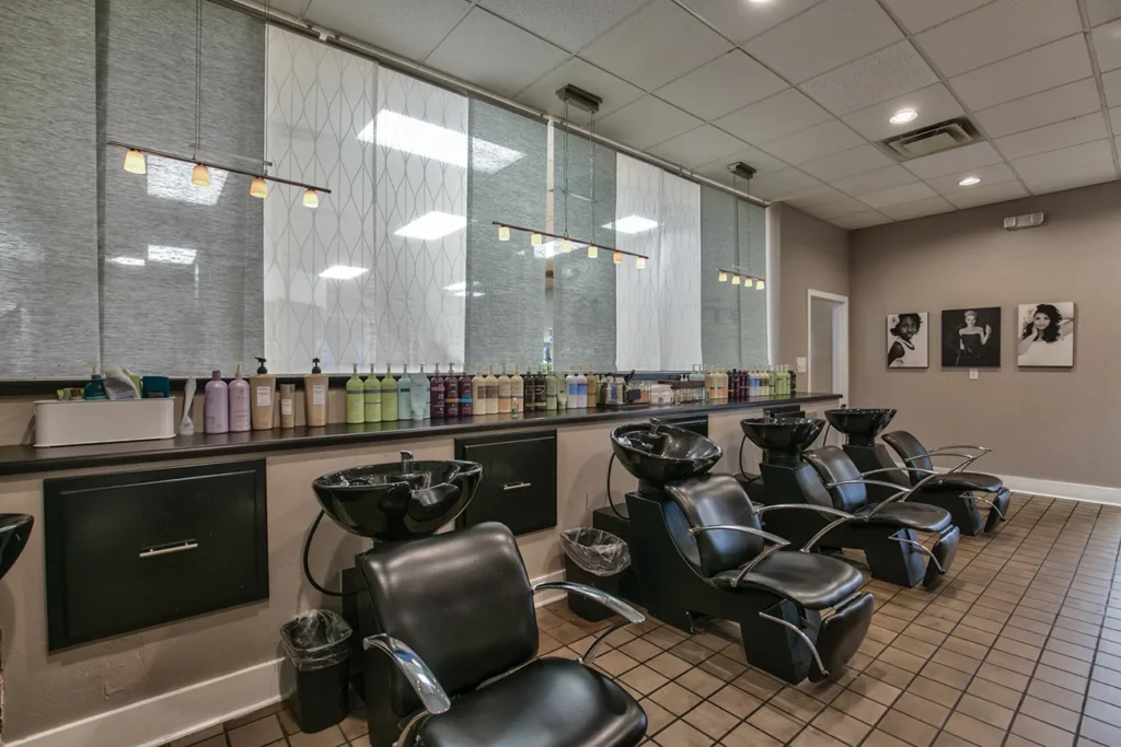 A row of black salon chairs and washbasins in a hair salon, with shelves of hair products on the wall and black-and-white portraits hanging in the background. - Soleil 7 Hair Salon in Tallahassee, FL
