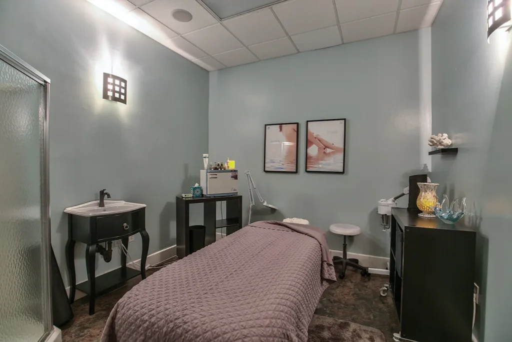 A small spa treatment room with a massage table, sink, counter, and decor, including two framed photos on a light blue wall. - Soleil 7 Hair Salon in Tallahassee, FL