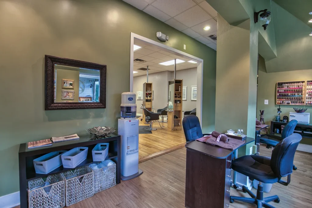 Modern salon interior with olive green walls, workstations, a water cooler, wall mirror, storage baskets, and manicure desk; shelves with nail polish are visible in the background. - Soleil 7 Hair Salon in Tallahassee, FL