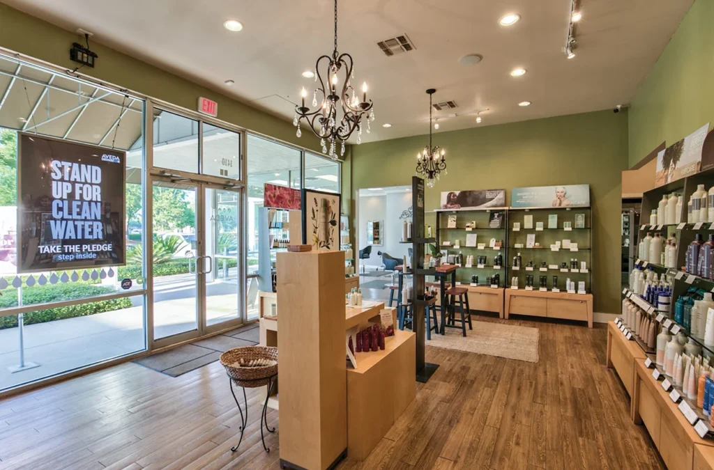 A modern salon interior with wood floors, product displays, shelves with hair products, chandeliers, and large windows near the entrance. A clean water awareness sign is visible by the door. - Soleil 7 Hair Salon in Tallahassee, FL