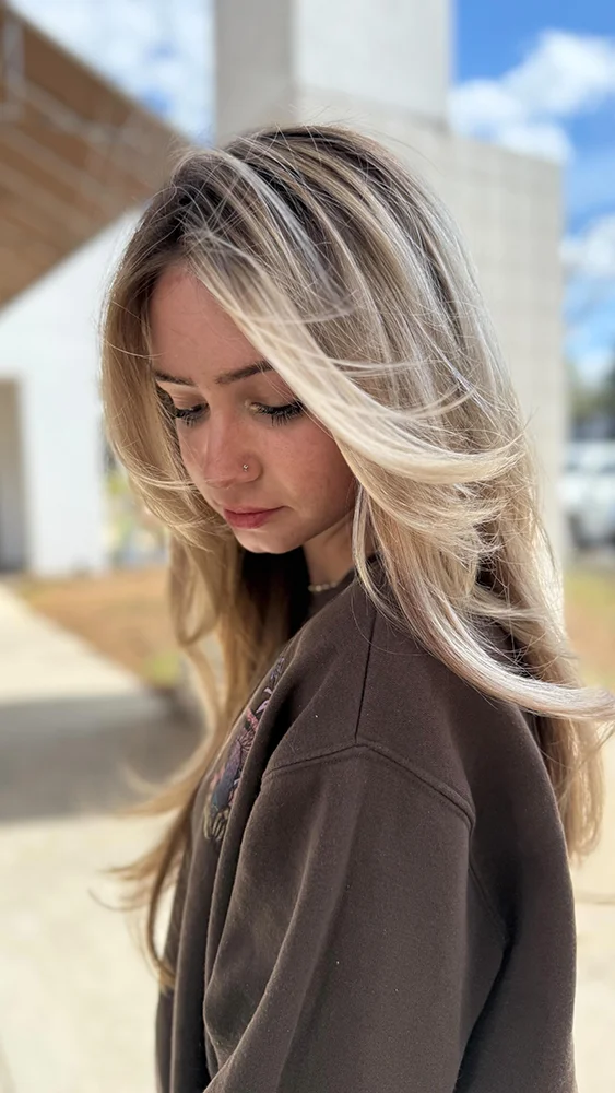 Young woman with long, layered blonde hair and a nose piercing looks down while standing outdoors in natural light. - Soleil 7 Hair Salon in Tallahassee, FL