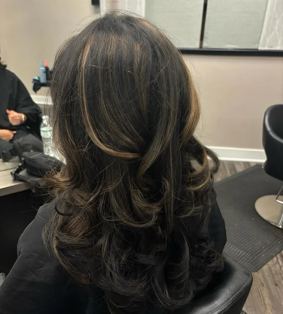 A person with long, dark, wavy hair featuring subtle caramel highlights sits in a salon chair, facing away from the camera. - Soleil 7 Hair Salon in Tallahassee, FL