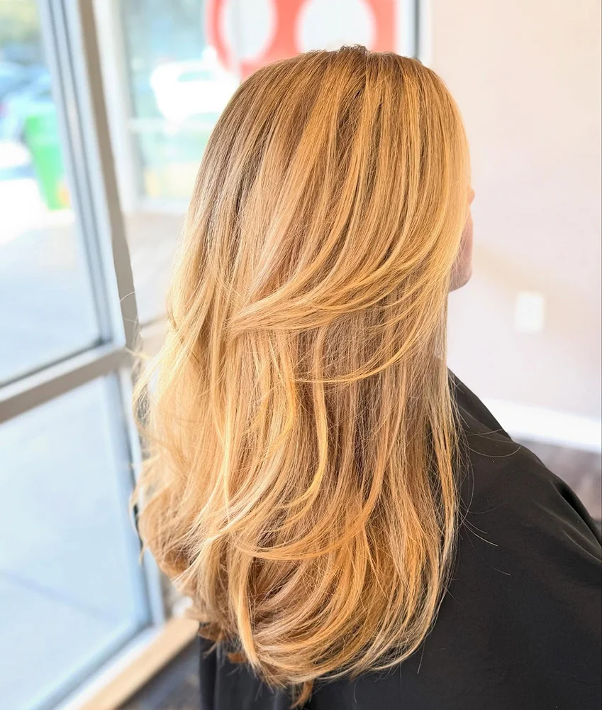 Person with long, layered, blonde hair styled with soft waves, shown from the back indoors near a window. - Soleil 7 Hair Salon in Tallahassee, FL