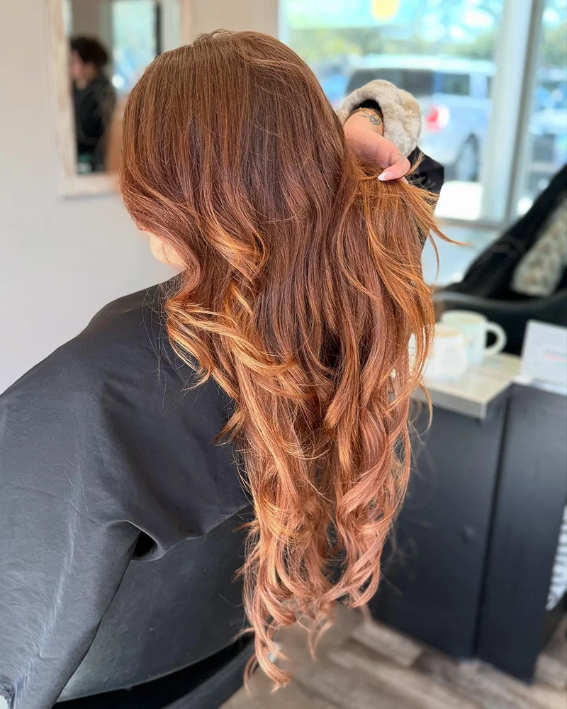 Person with long, wavy, reddish-brown hair is seated in a salon chair, with a stylist lifting a section of their hair. - Soleil 7 Hair Salon in Tallahassee, FL