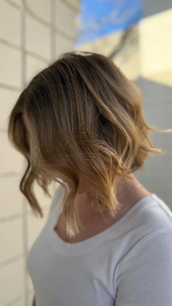 Person with wavy, shoulder-length light brown hair wearing a white top, turned slightly away from the camera, standing against a light-colored wall. - Soleil 7 Hair Salon in Tallahassee, FL