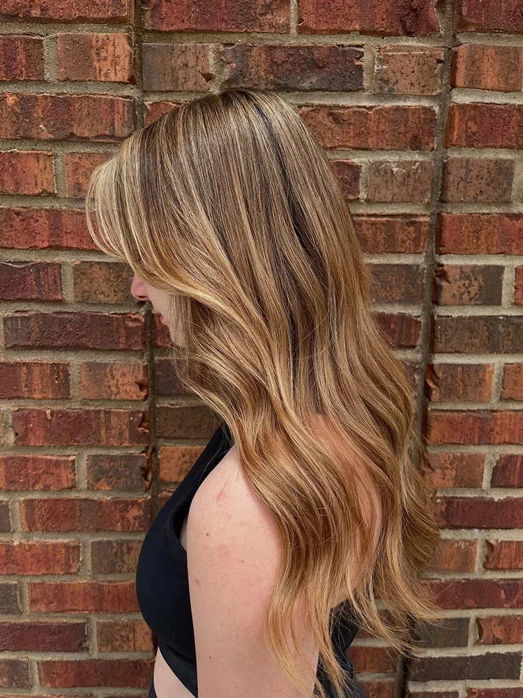 Person with long, wavy, blonde-highlighted hair standing sideways against a red brick wall, wearing a black sleeveless top. - Soleil 7 Hair Salon in Tallahassee, FL