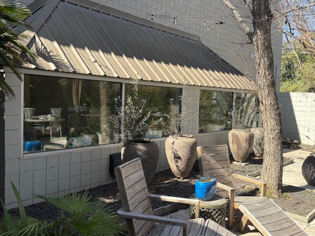 Outdoor patio with wooden chairs, large ceramic planters, a small blue table, and bare trees beside a building with large windows and beige square tiles. - Soleil 7 Hair Salon in Tallahassee, FL