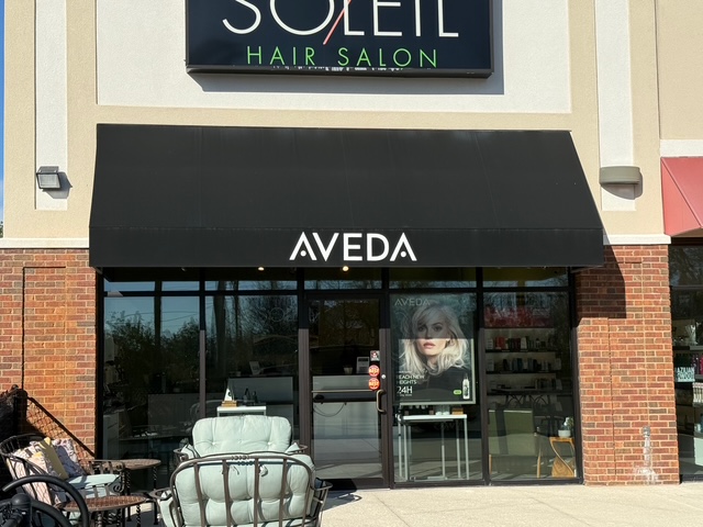 Storefront of Soleil Hair Salon with a black awning displaying "AVEDA." Outdoor seating is in front, and hair products are visible through large windows. - Soleil 7 Hair Salon in Tallahassee, FL
