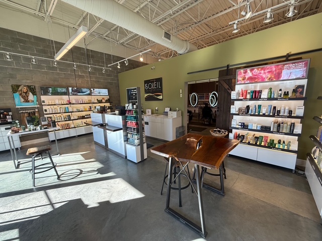 A modern salon interior with display shelves of hair and beauty products, a wooden table in the center, and sunlight streaming through large windows. - Soleil 7 Hair Salon in Tallahassee, FL
