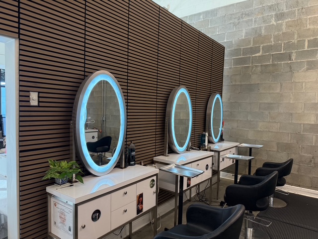Three salon stations with round, illuminated mirrors, white cabinets, and black chairs are arranged against a wood-paneled and concrete wall. - Soleil 7 Hair Salon in Tallahassee, FL