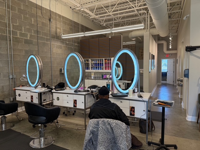 A person sits in a modern hair salon with circular lighted mirrors, styling stations, and shelves stocked with hair products. - Soleil 7 Hair Salon in Tallahassee, FL