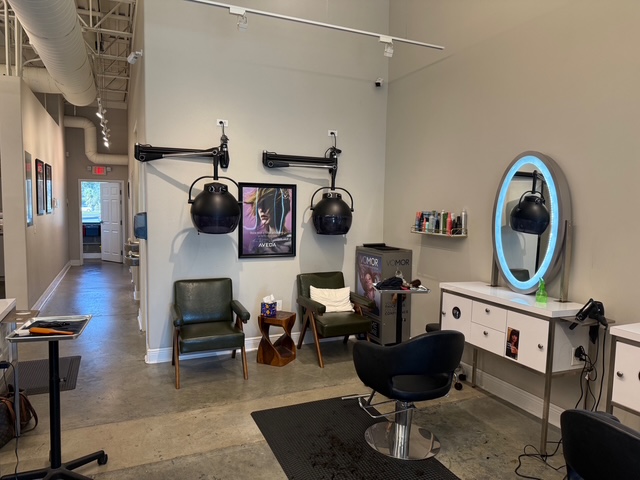 A modern hair salon with two green chairs under wall-mounted hair dryers, a black styling chair, a lighted mirror, and shelves with hair products. - Soleil 7 Hair Salon in Tallahassee, FL
