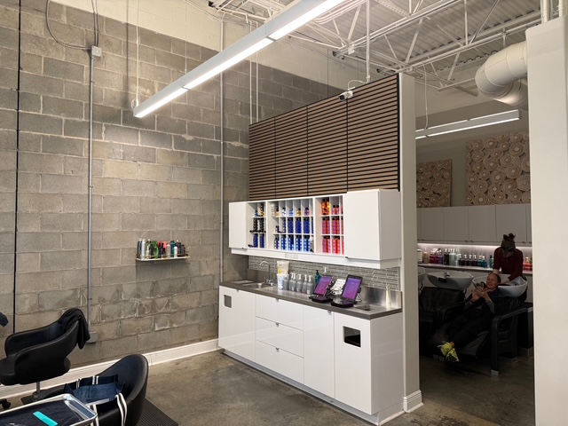 Modern salon interior with concrete walls, white cabinetry, hair products on display, and two people seated at wash stations in the background. - Soleil 7 Hair Salon in Tallahassee, FL