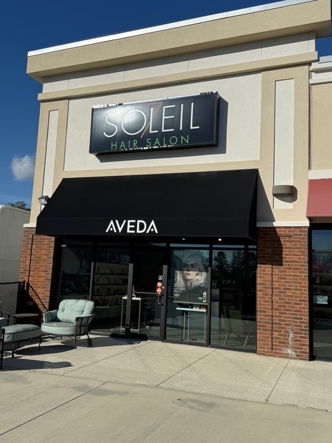 Exterior of Soleil Hair Salon with an Aveda sign, black awning, glass doors, a poster, and outdoor seating on a sunny day. - Soleil 7 Hair Salon in Tallahassee, FL
