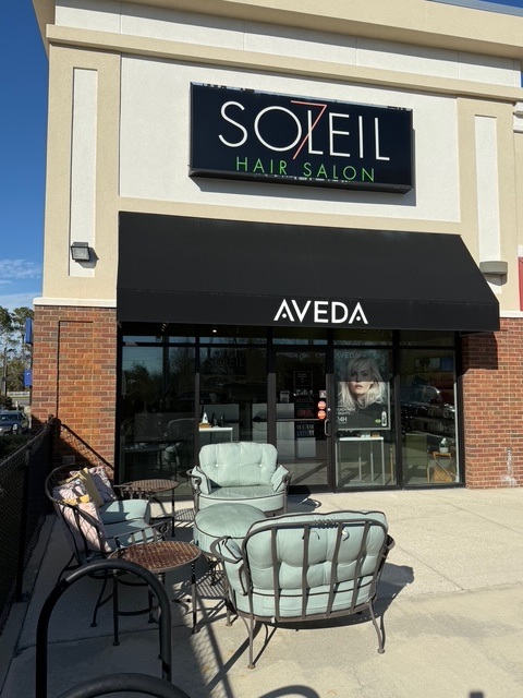 Exterior of Soleil Hair Salon with a black awning labeled "Aveda," glass door, large window, and outdoor seating area with cushioned chairs and a table. - Soleil 7 Hair Salon in Tallahassee, FL