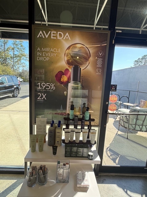 A display of Aveda hair and skin products is set up in front of a window, with a promotional poster for a serum behind it. - Soleil 7 Hair Salon in Tallahassee, FL