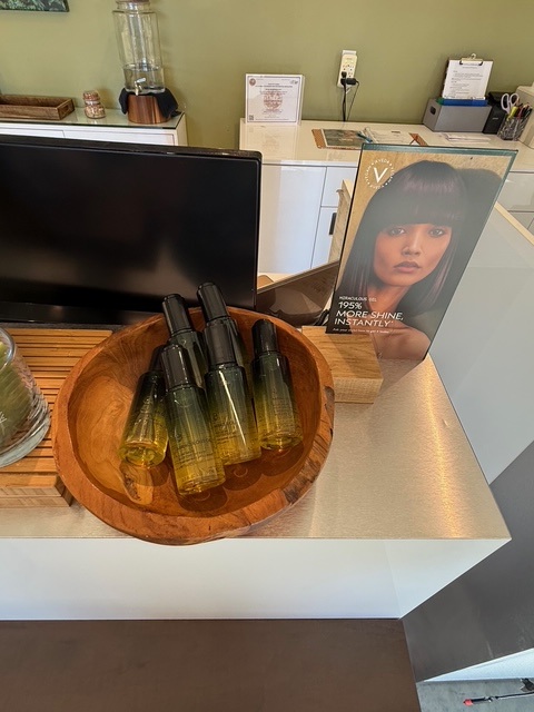 A wooden bowl holding several bottles of hair product displayed on a counter next to a promotional stand with a woman’s photo and product information. - Soleil 7 Hair Salon in Tallahassee, FL