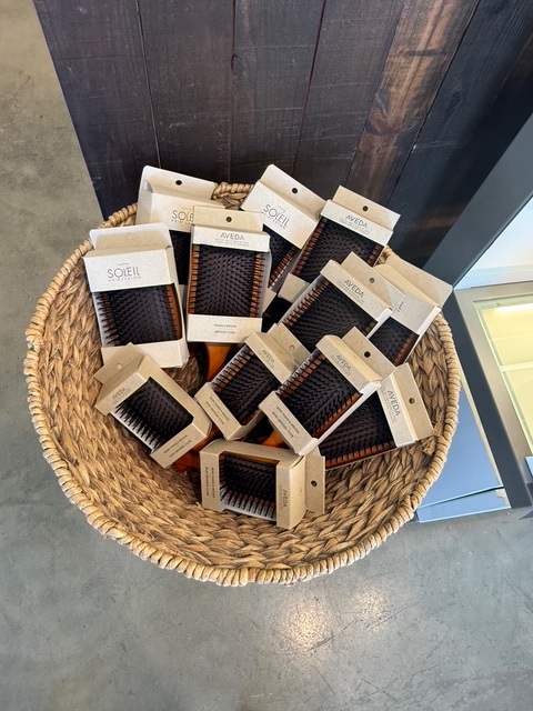 A wicker basket filled with multiple packaged wooden hairbrushes labeled "Aveda" and "Soleil" placed on a concrete floor. - Soleil 7 Hair Salon in Tallahassee, FL