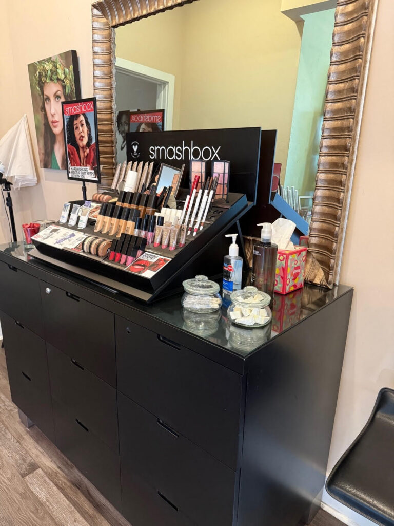 A black makeup station holds a Smashbox cosmetics display, tissues, hand sanitizer, jars, and a mirror. Posters and beauty products are visible in the background. - Soleil 7 Hair Salon in Tallahassee, FL