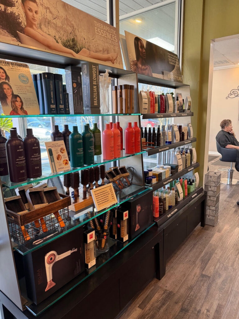 Shelves in a salon displaying various hair care products, including shampoos, conditioners, styling tools, and promotional signs, with a woman seated in the background. - Soleil 7 Hair Salon in Tallahassee, FL