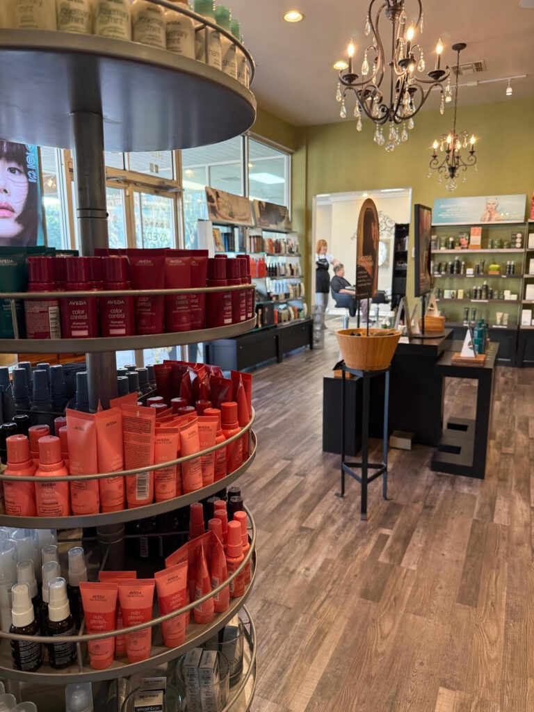 A salon retail area with shelves of hair care products, a chandelier overhead, wooden flooring, and a person visible in the background near salon chairs. - Soleil 7 Hair Salon in Tallahassee, FL