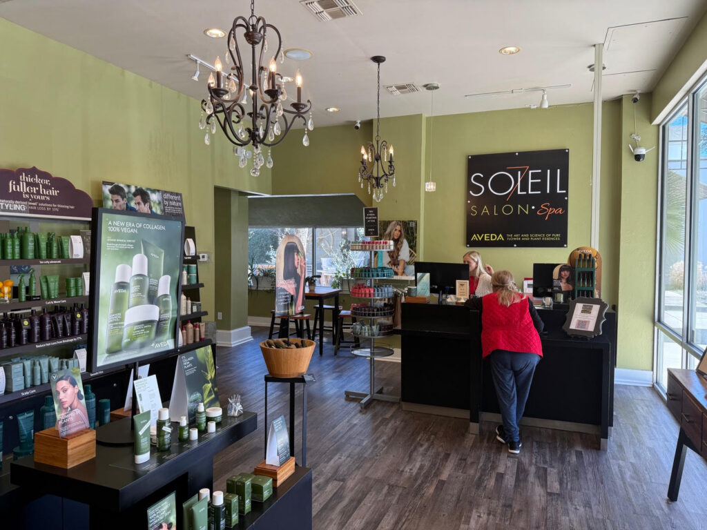 Interior of Soleil Salon & Spa showing the reception desk, hair products on display, two people interacting at the counter, and large windows letting in natural light. - Soleil 7 Hair Salon in Tallahassee, FL