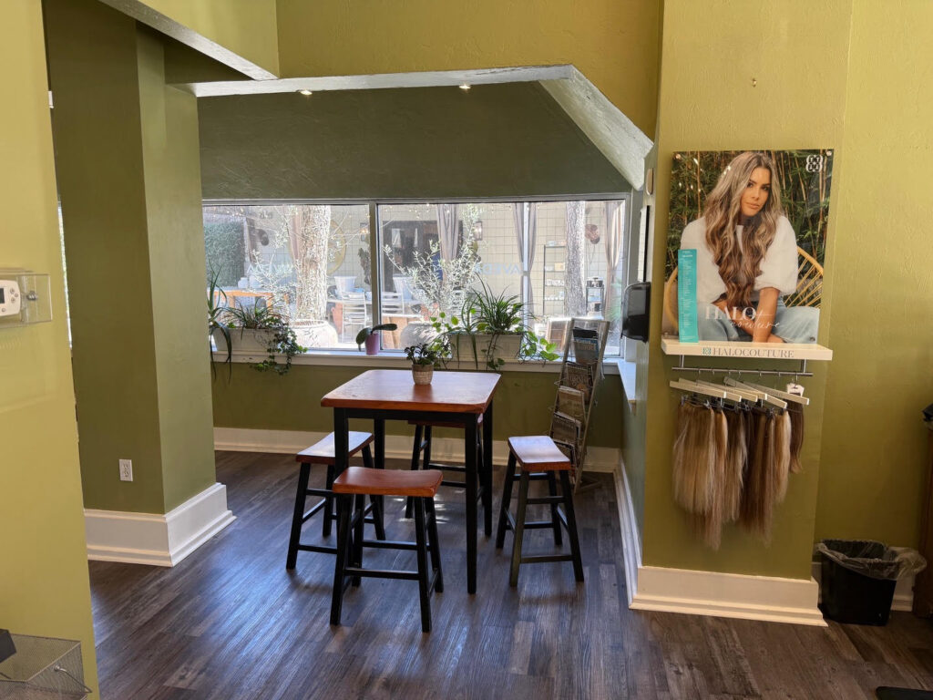 A small wooden table with four stools is placed by large windows in a room with green walls and dark wood flooring. A display of hair extensions and a poster are on the right wall. - Soleil 7 Hair Salon in Tallahassee, FL