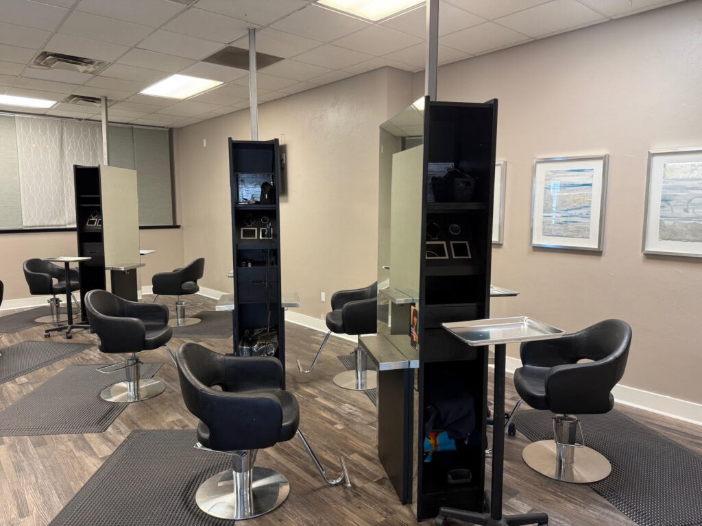A modern hair salon with black styling chairs, mirrors, and workstations arranged in rows. The floor is dark wood, and artwork is displayed on light-colored walls. - Soleil 7 Hair Salon in Tallahassee, FL