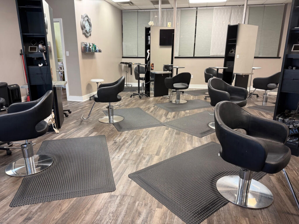 A modern hair salon with several black styling chairs, mirrors, and floor mats, set on wood-patterned flooring with neutral-colored walls. - Soleil 7 Hair Salon in Tallahassee, FL