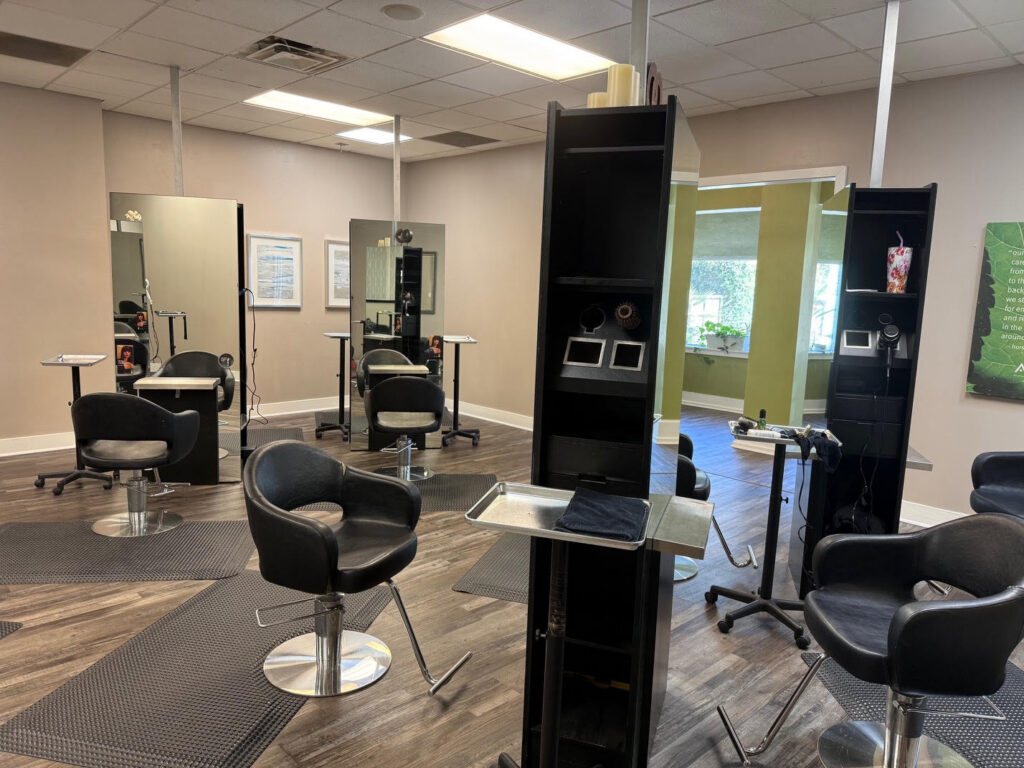 A modern hair salon interior with several black styling chairs, mirrors, and workstations on a brown wood floor. The space is clean and well-lit with natural and artificial light. - Soleil 7 Hair Salon in Tallahassee, FL