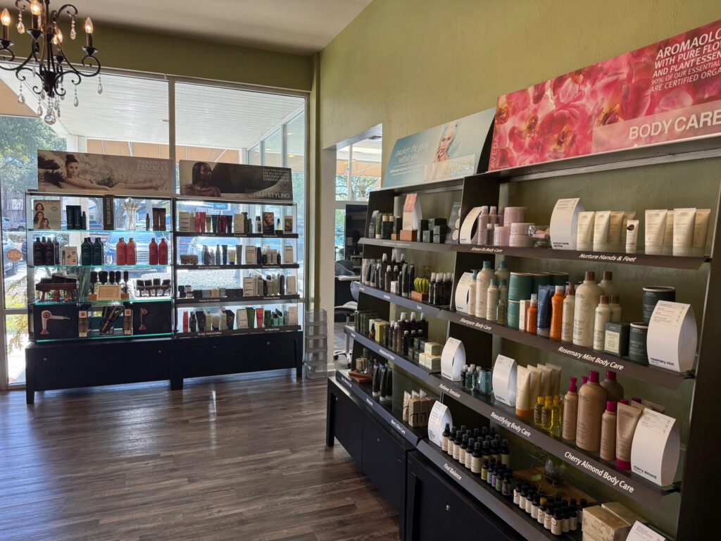 Shelves in a brightly lit store display various hair and body care products in bottles and containers, with large windows letting in natural light. - Soleil 7 Hair Salon in Tallahassee, FL