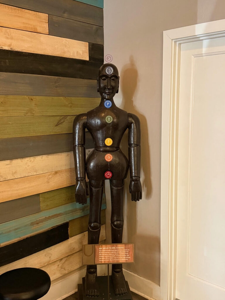 A wooden human figure stands upright, displaying seven chakra symbols along its body, positioned against a multicolored wood-paneled wall near a white door. - Soleil 7 Hair Salon in Tallahassee, FL