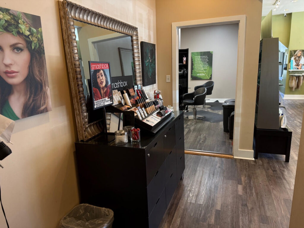 A salon interior with a makeup display, large mirror, wall art, salon chairs, and wood flooring. A makeup magazine is visible on the vanity. - Soleil 7 Hair Salon in Tallahassee, FL