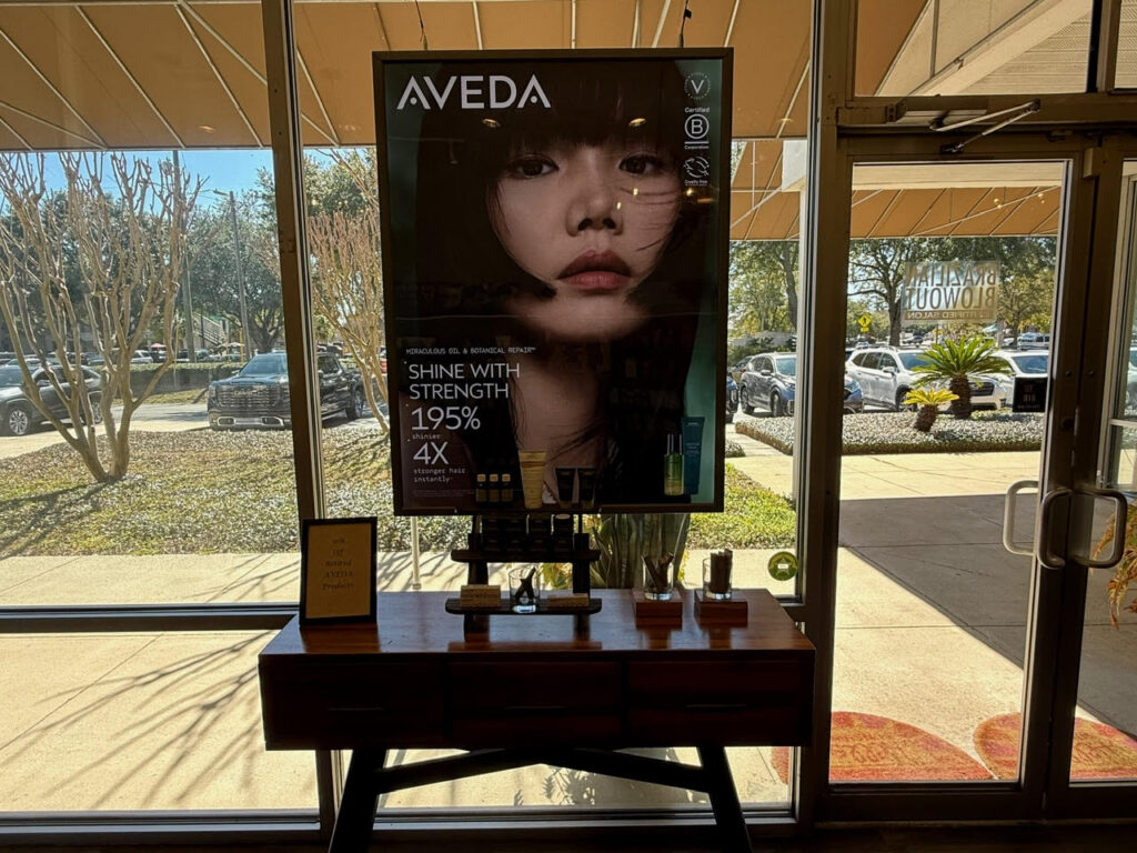 A promotional Aveda poster featuring a woman's face is displayed above a wooden table with hair products in a sunlit store entrance. - Soleil 7 Hair Salon in Tallahassee, FL