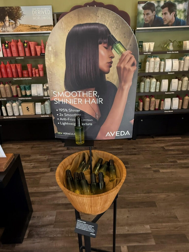 A display for Aveda hair products features a sign with a woman applying hair serum and a bowl below containing sample bottles, set in a store with shelves of hair care products in the background. - Soleil 7 Hair Salon in Tallahassee, FL