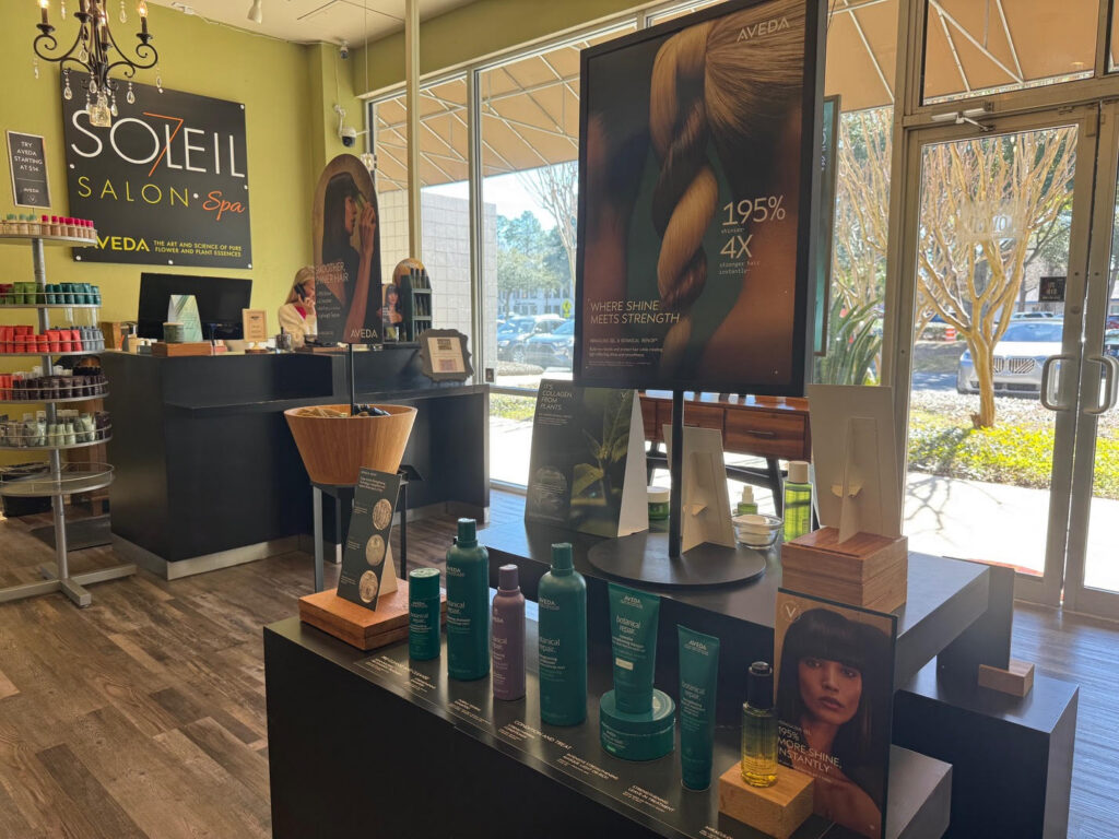 A salon reception area with hair products on display, promotional posters, and a front desk labeled “Soleil Salon Spa” with large windows letting in natural light. - Soleil 7 Hair Salon in Tallahassee, FL