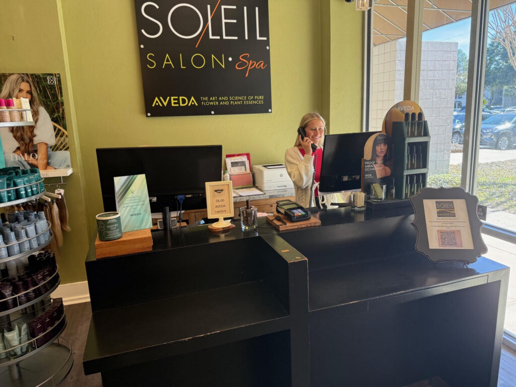 A woman sits at a salon reception desk, speaking on the phone. The counter displays products, signage, and a computer. A Soleil Salon + Spa sign hangs on the wall behind her. - Soleil 7 Hair Salon in Tallahassee, FL