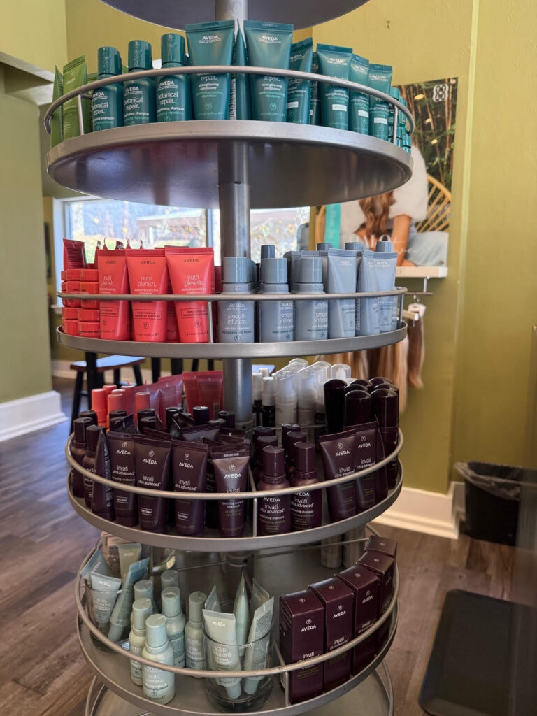 Rotating display shelf holding neatly organized Aveda hair and skincare products in tubes and bottles, sorted by color, inside a salon with green walls and wooden floors. - Soleil 7 Hair Salon in Tallahassee, FL