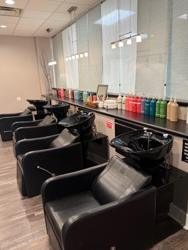 Three black salon shampoo chairs with sinks are positioned in front of a counter lined with colorful hair product bottles in a well-lit salon. - Soleil 7 Hair Salon in Tallahassee, FL