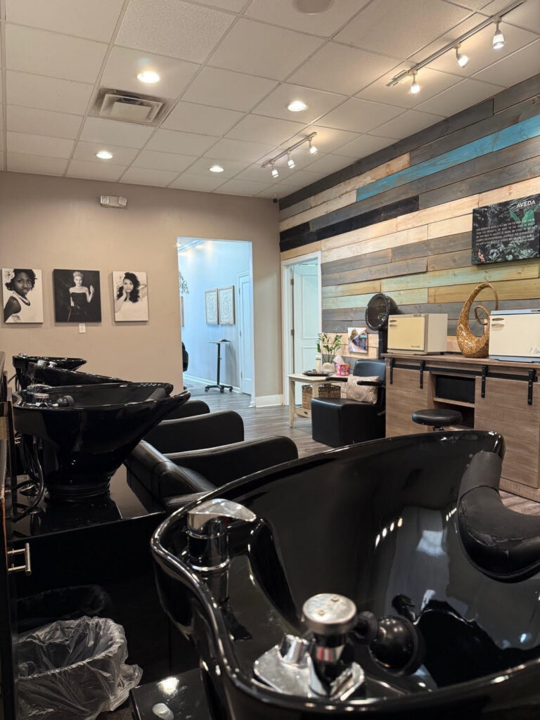 A modern hair salon interior with black wash stations, a textured wood accent wall, and framed portraits on a beige wall. The space is well-lit with ceiling lights. - Soleil 7 Hair Salon in Tallahassee, FL